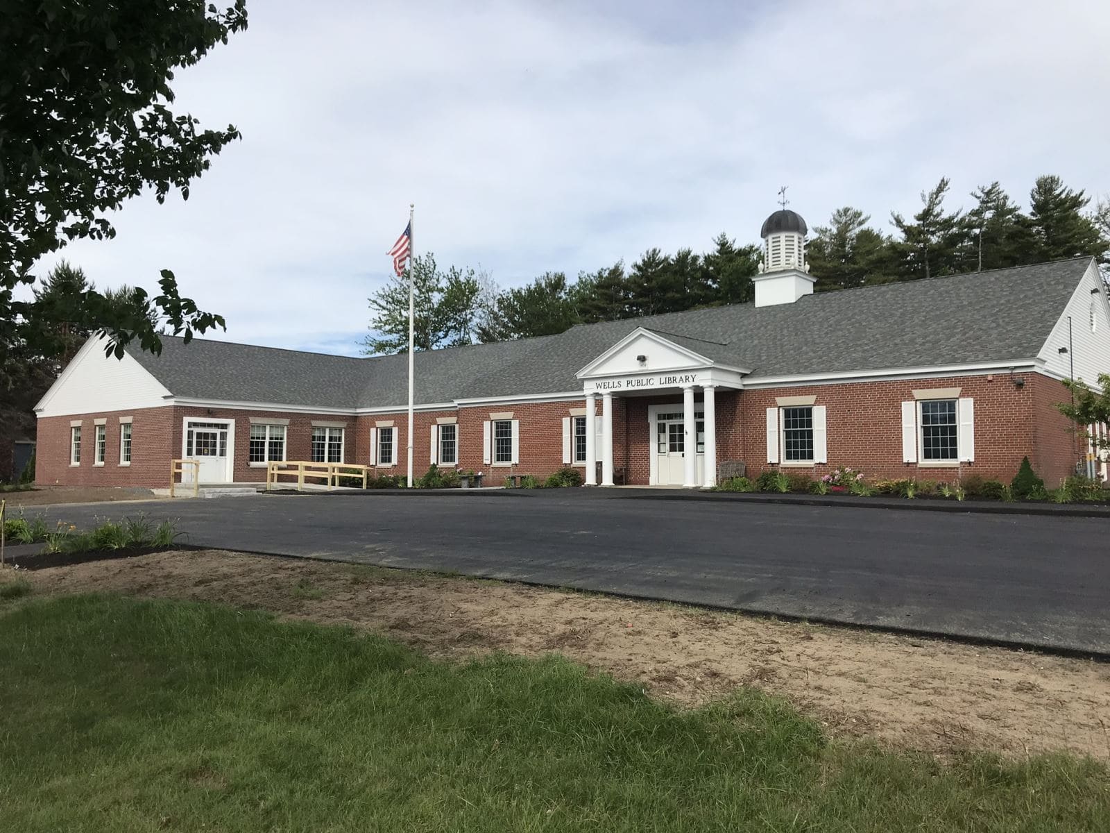 A red-brick building labeled Wells Public Library with white trim, a cupola, and an American flag out front, surrounded by greenery and trees under a cloudy sky.