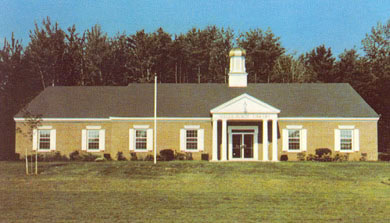 A single-story brick building with white trim, columns at the entrance, and a small cupola on the roof, set on a grassy lawn with trees in the background under a clear sky.
