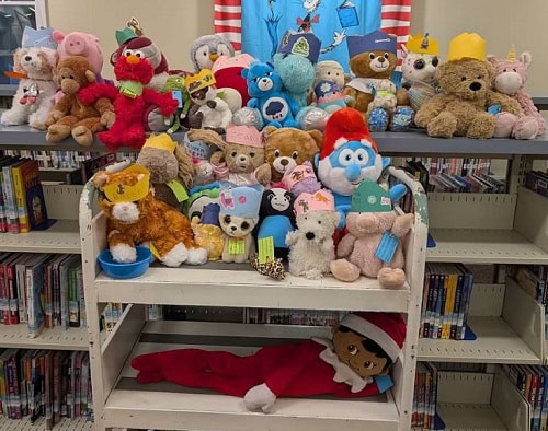 A display of stuffed animals, many wearing paper crowns, is arranged on white shelves in front of bookshelves. An Elf on the Shelf doll in red lies on the bottom shelf. A blue Dr. Seuss poster hangs behind the display.